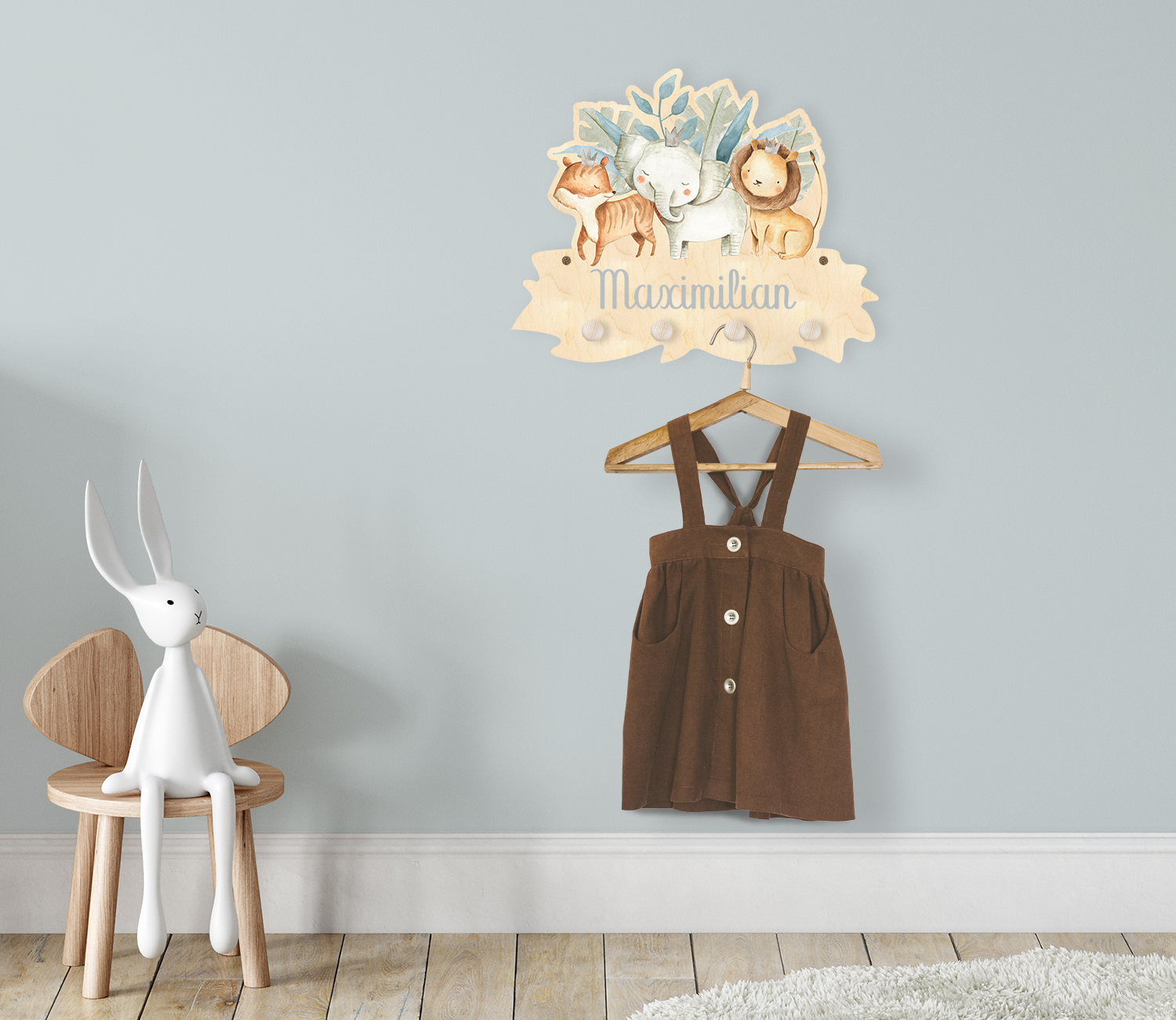 Die Kindergarderobe aus Holz mit augelasertem Namen und den Tieren: Tiger, Elefant und Löwe im Dschungel ist montiert an einer Kinderzimmerwand. An der Garderobe hängt ein Kleiderhaken mit einem Kleidungsstück.